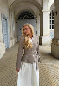 Femme blonde en veste beige à taille nouée et jupe blanche se tient sous une arche en pierre avec de hautes colonnes et des portes grises.