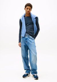 Junger Mann steht mit einer Hand in der Tasche, trägt ein marineblaues langärmeliges Hemd, einen blauen Strickpullover über den Schultern, lässige Jeans und Turnschuhe.