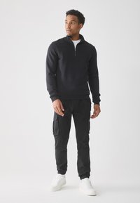 Zwarte zip-up sweatshirt met een hoge kraag, gecombineerd met zwarte cargo broek met zijzakken, en witte sneakers.