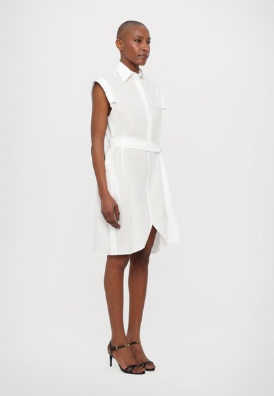 White shirt dress with short sleeves, button front, and a belted waist. Smooth fabric with a knee-length cut. Worn with black strappy heels.