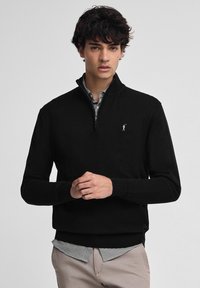 Ung man som bär en svart tröja med kvart zip över en svartvit rutad skjorta och beige byxor, stående mot en neutral bakgrund.