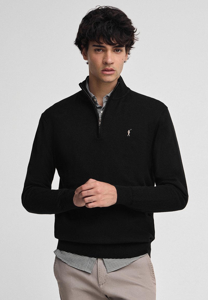 Ung man som bär en svart tröja med kvart zip över en svartvit rutad skjorta och beige byxor, stående mot en neutral bakgrund.