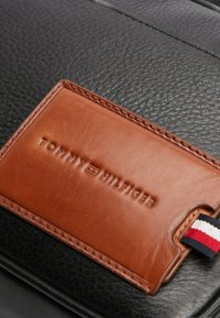 Borsa in pelle nera con etichetta in pelle marrone con nome del brand impressa, arricchita da un dettaglio in tessuto a strisce rosse, bianche e blu.