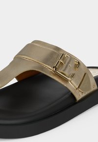 Μαύρη σαγιονάρα με φαρδιά μεταλλική χρυσή λωρίδα και λεπτομέρεια χρυσής αγκράφας στο πάνω μέρος του ποδιού.