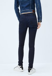 Marina smala jeans med hög midja, med bakfickor och subtila sömnadsdetaljer, tillsammans med vita sneakers och en jeansjacka.