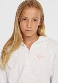 Hvid hættejakke med en blød tekstur og hjerte-logo i lys pink. Har hætte og frontlynlås, båret over en pinkstribet skjorte.