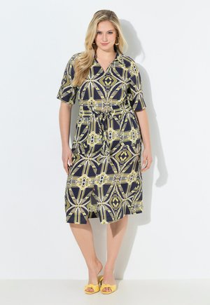 Kobieta stoi przy białej ścianie, ubrana w czarno-żółtą midi sukienkę w wzory z krótkimi rękawami oraz żółte buty na obcasie z odkrytymi palcami.