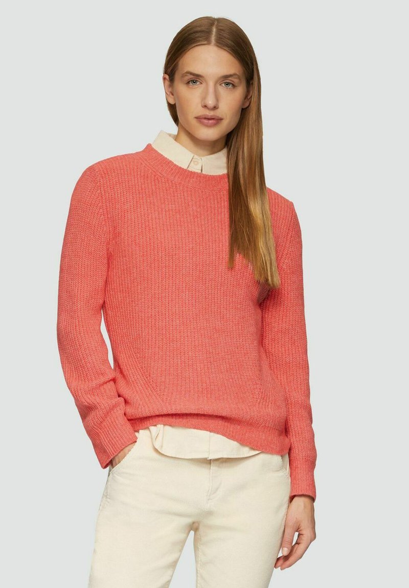 Korallenfarbener Pullover mit gerippter Textur, Rundhalsausschnitt und langen Ärmeln, über einem cremefarbenen Kragenhemd getragen. Über cremefarbenen Hosen.