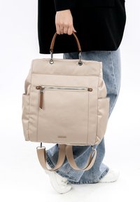 Person hält einen beigen Rucksack mit braunen Griffen und einer vorderen Reißverschlusstasche, trägt blaue Jeans, weiße Turnschuhe und eine schwarze Jacke.