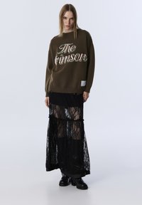 Olivgrüner, gestrickter Pullover mit "The Crimson" in bunten Buchstaben gestickt, kombiniert mit einem geschichteten, schwarzen Spitzen-Maxirat und klobigen schwarzen Stiefeln.