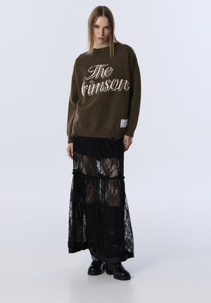 Olivově zelený pletený svetr s vyšitým nápisem "The Crimson" v barevných písmenech, zkombinovaný s vrstvenou černou krajkovou maxi sukni a robustními černými botami.