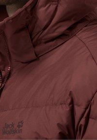 Veste isolante bordeaux avec col haut, finition lisse, fermeture éclair à l'avant et logo "Jack Wolfskin" en bas à gauche.