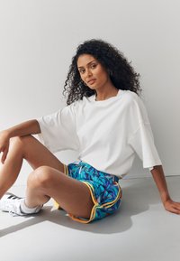 Maglietta bianca oversize abbinata a pantaloncini colorati con motivo multicolore su sfondo blu, foglie tropicali e rifiniture gialle. Scarpe da ginnastica bianche.