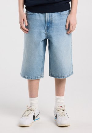 Person trägt hellblaue, kniehohe Jeans-Shorts, weiße Sportsocken und weiße Nike-Sneaker mit schwarzem Swoosh.