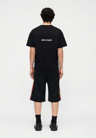 Άνδρας που στέκεται με την πλάτη προς το μέρος του θεατή, φοράει μαύρο μπλουζάκι "Palm Angels", μαύρα σορτς με λωρίδες στα πλάγια, μαύρες κάλτσες και μαύρα αθλητικά παπούτσια.