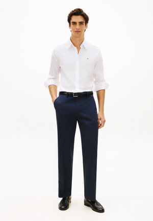 Biała koszula na guziki, granatowe spodnie, czarny pasek i czarne buty. Koszula ma długie rękawy z podwiniętymi mankietami i małe logo.