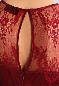 Burgundy lace garment featuring floral patterns, sheer texture, and a keyhole back with a button closure. Detailed stitching on edges.