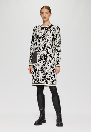 Robe longueur genoux de couleur noire avec un motif floral blanc et bleu pâle, dotée de manches longues et d'une poche avant. Accents noirs unis au niveau de l'ourlet.
