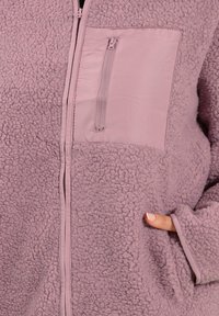 Chaqueta de forro polar en malva claro con un cuerpo texturizado. Presenta un bolsillo de tela suave y cremallera, con bolsillos laterales y puños elásticos.