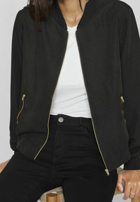 Zwarte bomberjack met een gladde afwerking, gouden ritssluitingen, geribde kraag en zijzakken. Draagt over een wit shirt en zwarte jeans.