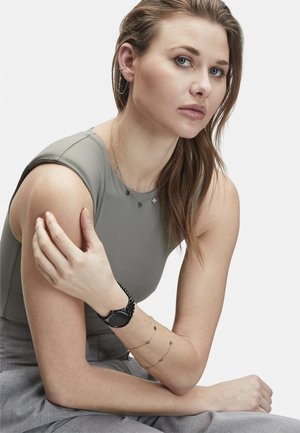 Schwarze Uhr mit strukturiertem Armband, geschichtete silberne Halsketten, graues ärmelloses Oberteil und hellgraue Hose. Warmer Hautton, natürliches Make-up, lange Haare.