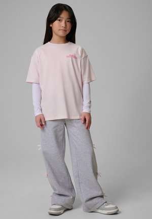 Młoda osoba stoi, ubrana w jasnoróżowy oversize t-shirt nałożony na białą koszulę z długim rękawem, szare szerokie spodnie z różowymi troczkami i szare trampki.