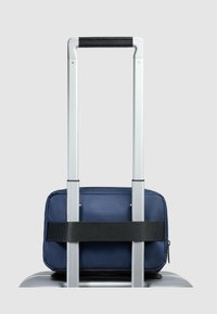 Borsa da viaggio blu navy con superficie testurizzata, fissata alla maniglia di una valigia argentata da una cinghia nera, forma rettangolare, design compatto.