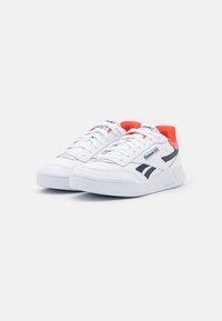 Paire de baskets Reebok blanches avec des bandes noires et une doublure intérieure rouge, présentées sur un fond blanc inclinées vers la droite.