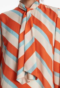 Langärmlige Bluse mit vertikalen diagonalen Streifen in Orange, Hellblau und Creme, mit einem Bindeband am Hals und weichem Stoffgefühl.