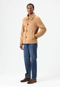 Tan faux shearling jas met knopen aan de voorkant, grote kraag en zijzakken, gecombineerd met blauwe spijkerbroek en bruine leren schoenen.