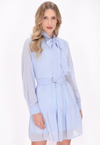 Vaaleansininen mekko, jossa on pilkullinen tekstuuri, kragenauha, pitkät läpinäkyvät hihat, vyötäröllä vyö hopeisella renkaalla ja kerroksellinen röyhelöhelma.