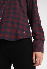Červeno-námořnická kostkovaná flanelová košile s dlouhými rukávy, knoflíková; má kulatý lem a malý stříbrný logo akcent na boku.