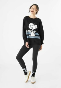 Sweatshirt noir avec un motif Snoopy, rayures bleues ; associé à des leggings noirs et des baskets blanches. Possède des cheveux bouclés et des chaussettes ludiques.