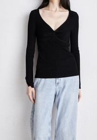 Czarny top z długim rękawem, głębokim dekoltem w kształcie litery V oraz marszczeniem na środku; prążkowana faktura; zestawiony z jasnoniebieskimi, szerokimi spodniami typu jeans.