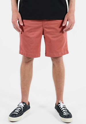 Homme portant un short rouille arrivant aux genoux, des baskets noires avec des lacets blancs, et une chemise noire, debout devant un fond uni.