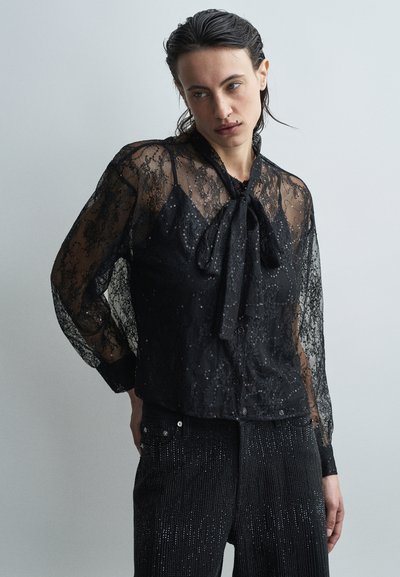 Blouse en dentelle noire avec des manches longues et un nœud au cou, ornée de subtils sequins. Associée à un pantalon noir orné.