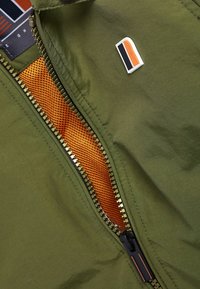 Grön jacka med en gyllene blixtlås, orange meshfoder och en liten logotyp-lapp med svarta och orange accenter. Strukturerad tygyta.