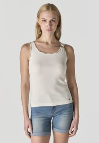 Plavolasa ženska z velikimi uhančki v obliki obročev, brezrokavnikom beige barve s podrobnostmi s kovicami in modrimi jeans kratkimi hlačami z zavihanimi robovi, stoji pred sivo podlago.