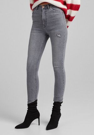 Jean gris slim avec légère usure à la cuisse, associé à des bottines chaussettes noires à talons hauts sur un fond clair.