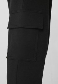 Černé cargo kalhoty z měkké tkaniny, které mají výraznou náprsní kapsu na horní části stehna a střih přiléhající k noze.