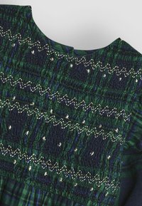 Kleid in Marineblau und Grün mit gemustertem Design, das eine gesmockte Textur, metallische Fäden und einen runden Halsausschnitt mit einem geplisséten Rockdesign aufweist.