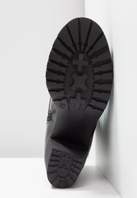 Semelle de botte noire montrant un motif de bande de roulement détaillé avec des formes en croix et des rainures, positionnée debout contre des murs d’angle blancs.