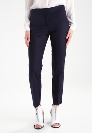 Granatowe zwężane spodnie wykonane z gładkiego materiału, z wysokim stanem i plisą z przodu, zestawione z białymi sandałami na obcasie z odkrytymi palcami.