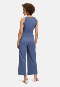 Combinaison bleue avec détail en volant aux épaules, taille cintrée et pantalons à jambes larges. Présente un fermeture à boutons au dos et une texture lisse.