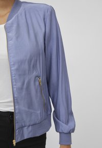 Ljuslila bomberjacka med en slät yta, gulddragkedja, ribbade muddar och sidoficka. Bärs över en vit topp.