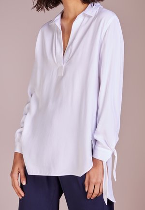 Femme portant un chemisier blanc ample à manches longues avec un décolleté en V profond et des détails à nouer aux poignets, associé à un pantalon foncé.