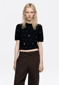 Suéter de punto negro de manga corta con adornos verdes y plateados. Combinado con pantalones marrones de cintura alta, creando un ajuste relajado.