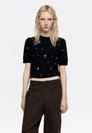 Suéter de punto negro de manga corta con adornos verdes y plateados. Combinado con pantalones marrones de cintura alta, creando un ajuste relajado.