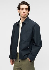 Μπλε σκούρο ελαφρύ μπουφάν με γιακά, μπροστινή τσέπη και λεπτομέρειες με κουμπιά, φορεμένο πάνω από λευκό πουκάμισο, συνδυασμένο με χακί παντελόνια.