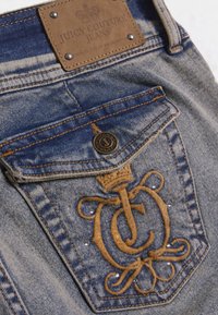 Τζιν παντελόνι με μπλε ξέπλυμα, καφέ δερμάτινη ετικέτα με ανάγλυφο λογότυπο και περίτεχνη καφέ κεντητική με διακοσμητικά στρας στην τσέπη.
